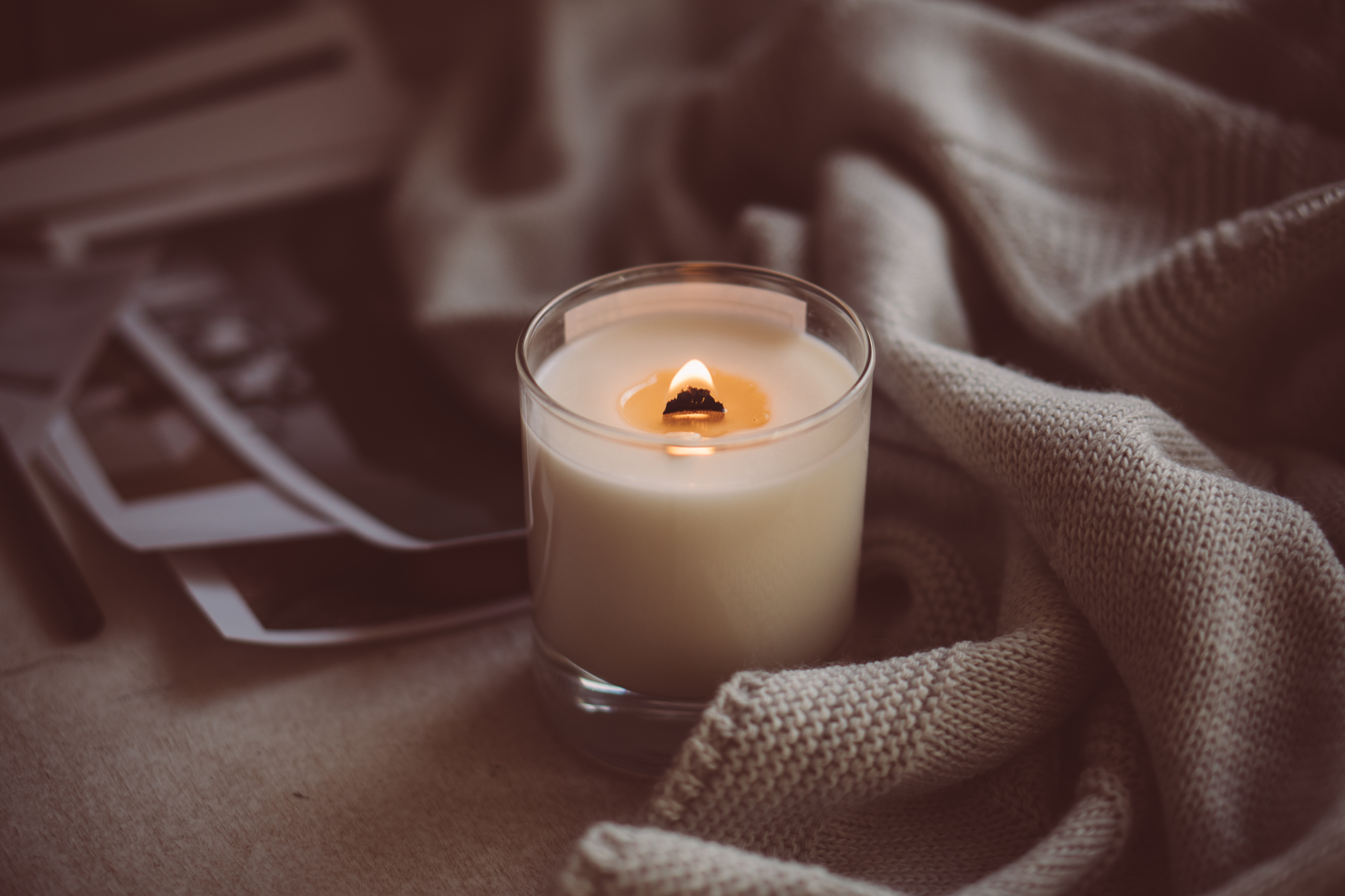A small candle glowing gently in a glass jar beside a soft knitted blanket in warm, low evening light at home