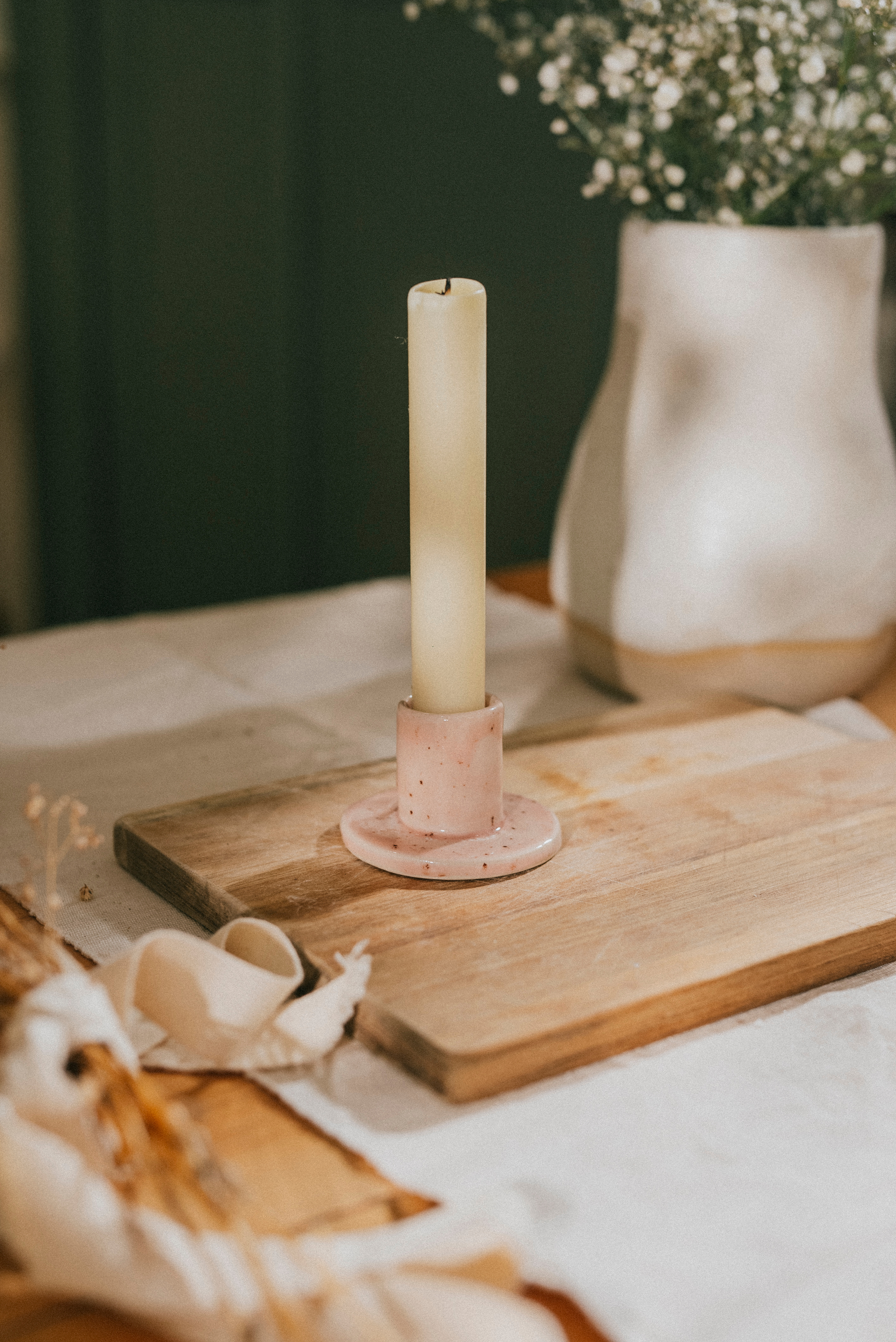 A single lit candle on a wooden table with soft flowers behind it, a tender and quiet corner of a home