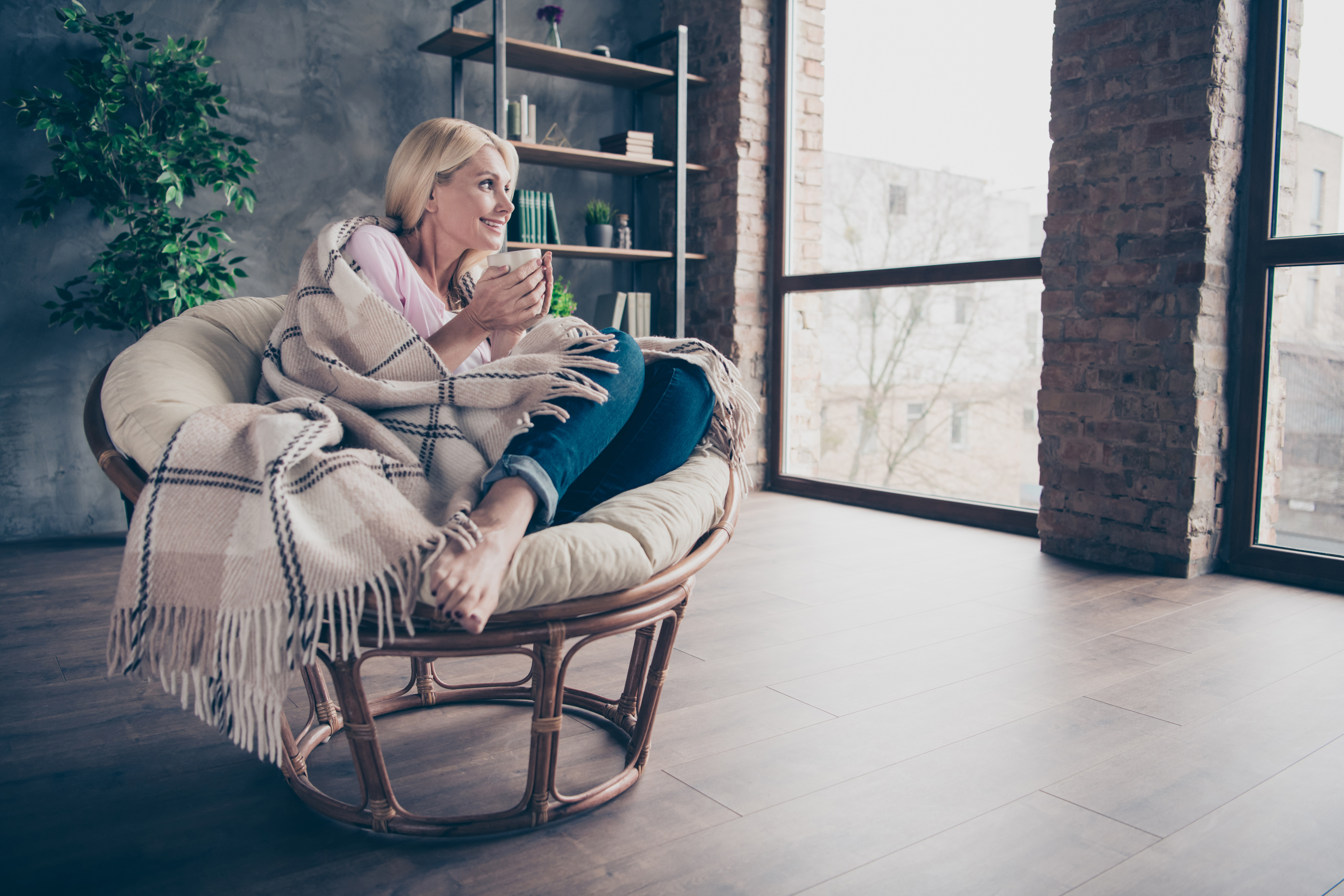 A woman wrapped in a checked blanket rests in an armchair with a mug, looking out a window on a quiet winter afternoon at home