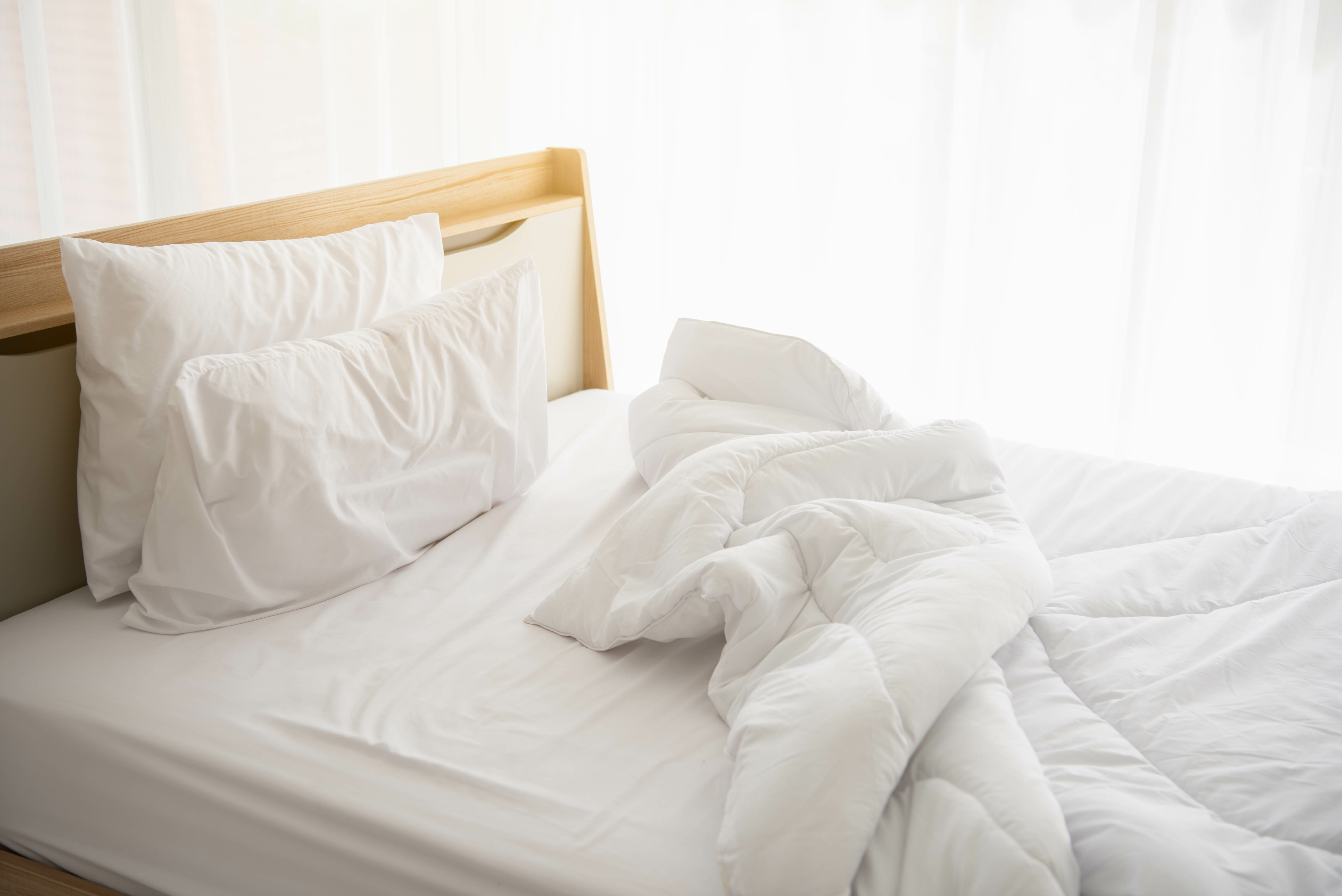Unmade white bed with rumpled linens beside a curtained window, filled with soft and forgiving morning light