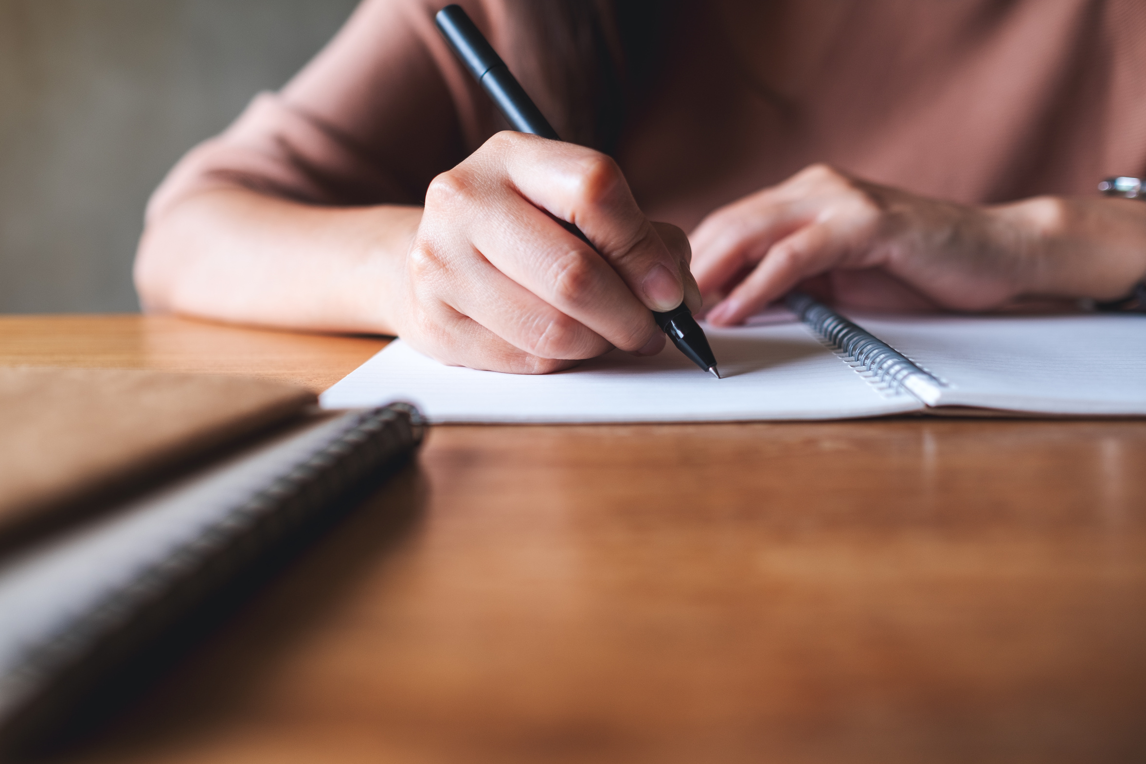 A woman's hands writing slowly in an open notebook at a wooden table, pen resting mid sentence, the unhurried pace of putting a few words down at a time