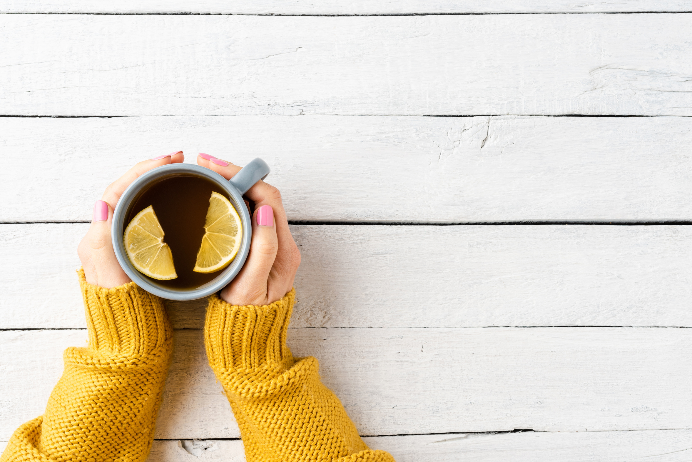A woman's hands in a soft knit sweater cradling a warm cup of tea at a pale wooden table, a small quiet comfort kept close during a hard season